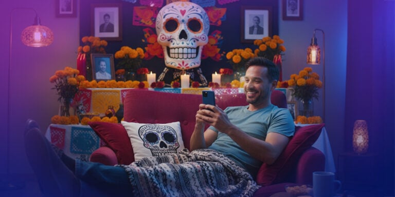 HOMBRE EN SU CASA DE ALREDEDOR 35 AÑOS LA CUAL ESTE SENTADO EN SU SILLON MIENTRAS TIENE UN PAN DE MUERTO DE FONDO TAMBIEN EL ESTA CON SU TELEFONO EN MANO ENTRETENIDO EN PLAYUZU